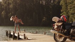 Молодий чоловік носить молоду жінку на причалі на березі озера біля мотоцикла — стокове фото