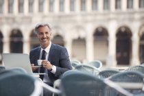 Hombre de negocios sonriente bebiendo espresso y usando el portátil en la cafetería de la acera - foto de stock