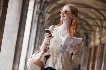 Усміхнена жінка тримає мобільний телефон і карту в коридорі — стокове фото