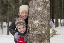 Portrait of happy boys behind tree trunk in snowy woods — Stock Photo