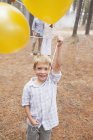 Портрет улыбающегося мальчика, держащего воздушные шары в лесу с родителями на заднем плане — стоковое фото