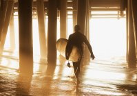 Älterer Surfer trägt Brett unter Pier — Stockfoto
