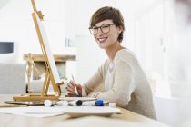 Портрет усміхненої жінки, що малює на мольберті на столі — стокове фото