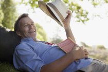 Portrait d'un homme souriant posé sur l'herbe avec un livre et un chapeau — Photo de stock