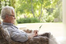Hombre mayor usando tableta digital en sillón de mimbre en el porche - foto de stock