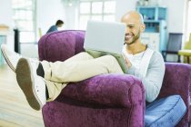 Young happy man using laptop in armchair — Stock Photo