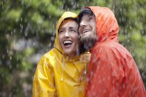 Casal feliz em capas impermeáveis olhando para a chuva — Fotografia de Stock