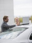 Улыбающийся бизнесмен и деловая женщина используют ноутбук на крыше автомобиля — стоковое фото