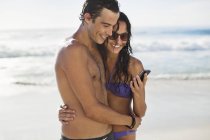Happy couple with cell phone hugging on beach — Stock Photo
