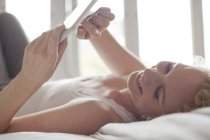 Mulher sorridente deitada na cama e usando tablet digital — Fotografia de Stock
