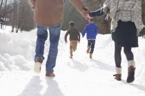 Вид сзади счастливой пары, держащейся за руки и смотрящей, как мальчики бегут по снежной полосе — стоковое фото