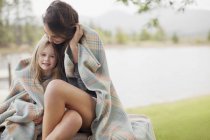 Retrato de hija sonriente envuelta en manta con madre a orillas del lago - foto de stock