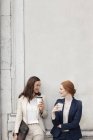 Smiling businesswomen drinking coffee and talking against building wall — Stock Photo