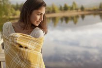 Улыбающаяся женщина, завернутая в одеяло на берегу озера — стоковое фото