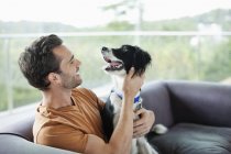 Усміхнений чоловік кидає собаку на диван в сучасному будинку — стокове фото