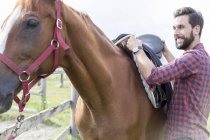 Lächelnder Mann entfernt Sattel vom Pferd — Stockfoto