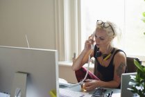Молода жінка з навушниками та татуюванням п'є чай і розмовляє по телефону в домашньому офісі — стокове фото