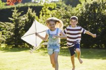 Hermano y hermana corriendo con cometa en jardín soleado - foto de stock