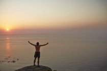 Exuberant male runner with arms outstretched on rock overlooking ocean sunset — Stock Photo