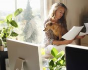 Femme avec chien examinant la paperasserie dans le bureau à domicile ensoleillé — Photo de stock
