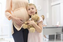 Porträt Mädchen mit Teddybär umarmt schwangere Mutter in Arztpraxis — Stockfoto