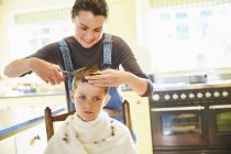 Unhappy boy getting haircut from mother in kitchen — Stock Photo