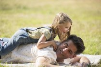 Playful daughter laying on top of father in sunny field — Stock Photo