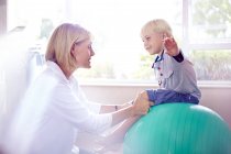 Physiothérapeute tenant garçon avec les bras tendus sur la balle de remise en forme — Photo de stock