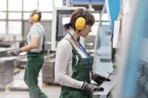 Arbeiterin mit Ohrenschutz hält Metallteil in Fabrik — Stockfoto