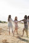 Mujeres boho cogidas de la mano y bailando en círculo en el soleado campo rural - foto de stock