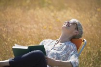 Mulher sênior lendo livro e rindo com a cabeça de volta no campo ensolarado — Fotografia de Stock