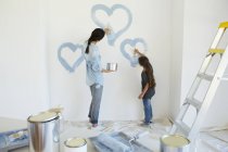 Madre e hija pintando corazones azules en la pared en una casa nueva - foto de stock