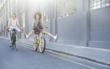 Donne giocose che costeggiano le biciclette lungo la strada urbana — Foto stock