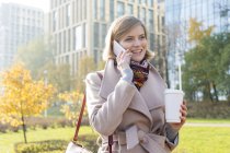 Усміхнена бізнес-леді з кавою розмовляє на мобільному телефоні в міському парку — стокове фото