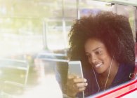 Mulher sorridente com afro ouvir música com fones de ouvido e mp3 player no ônibus — Fotografia de Stock