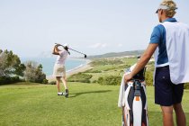 Caddy watching woman tee off on golf course overlooking ocean — Stock Photo