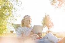Mulher sênior com fones de ouvido e vinho usando tablet digital na cadeira lounge no quintal ensolarado — Fotografia de Stock
