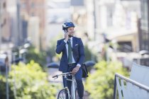 Businessman in suit and helmet sitting on bicycle talking on cell phone in city — Stock Photo
