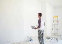 Человек с подносом с краской смотрит на белую стену — стоковое фото