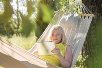 Senior woman using digital tablet and relaxing in summer hammock — Stock Photo