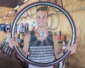 Retrato sonriente joven sosteniendo rueda de bicicleta en tienda de bicicletas - foto de stock