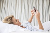 Femme plus âgée utilisant un téléphone portable sur le lit — Photo de stock