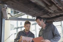Mechanics with clipboards talking under car in auto repair shop — Stock Photo