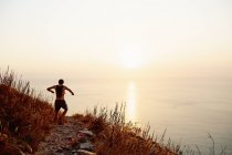 Мужчина бегун с рюкзаком спускается по тропинке с видом на закат океана — стоковое фото