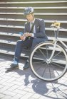 Businessman in suit and helmet texting with cell phone next to bicycle on sunny urban stairs — Stock Photo