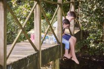 Brother and sister dangling bare feet over edge of footbridge — Stock Photo