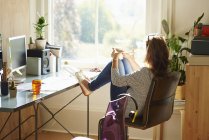 Mulher pensativa olhando através da janela com os pés na mesa no escritório ensolarado em casa — Fotografia de Stock