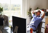 Человек откидывается назад, работая за компьютером в солнечном домашнем офисе — стоковое фото