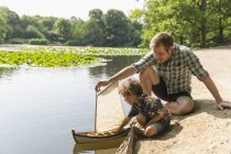 Pai e filho brincando com veleiro de brinquedo à beira do lago — Fotografia de Stock