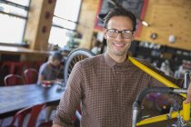 Portrait smiling man with eyeglasses carrying bicycle in cafe — Stock Photo